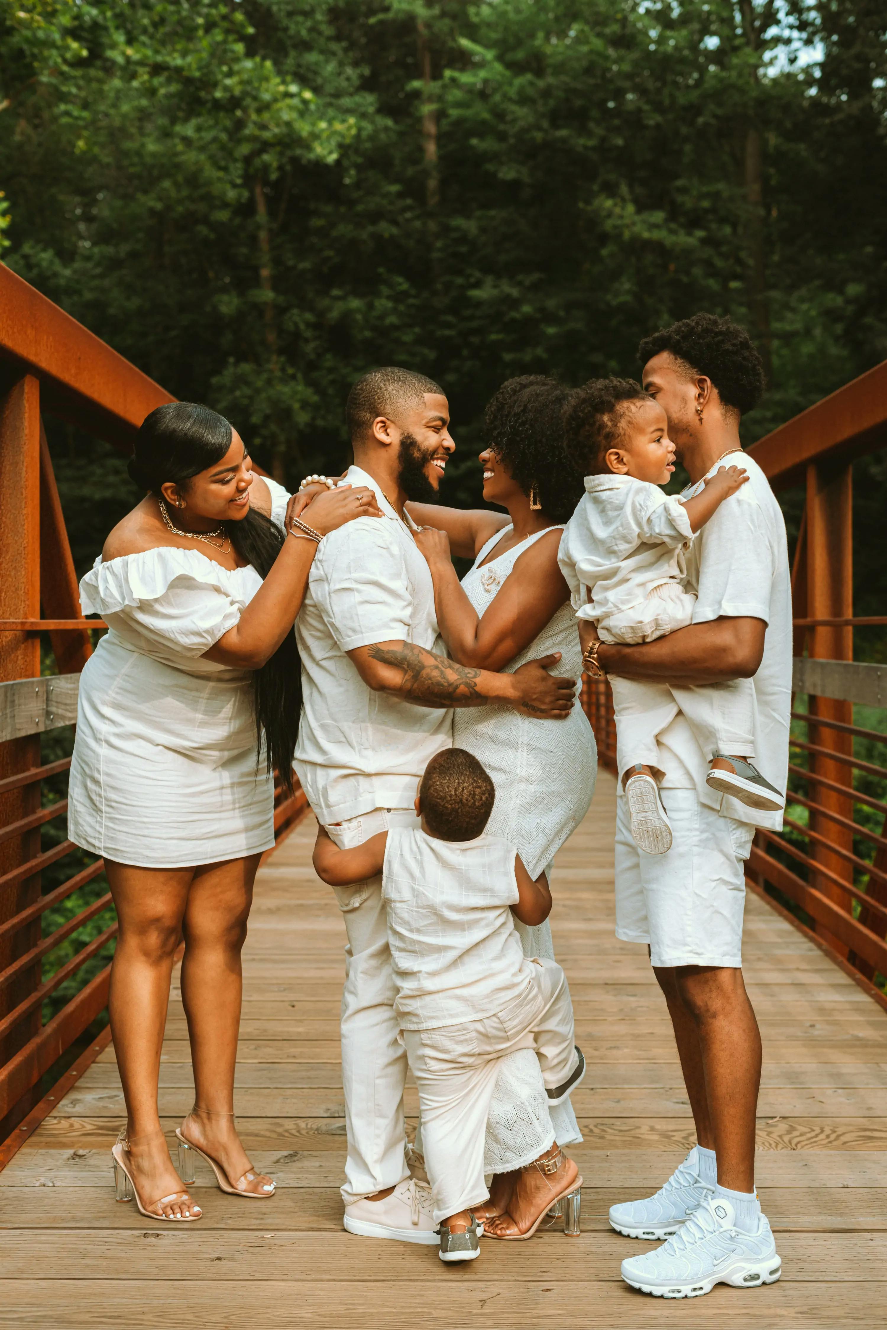Black mother and father smiling at each other in the middle of a footbridge, with oldest daughter on the left, oldest son on the right, holding their youngest son, and third son hugging their legs.